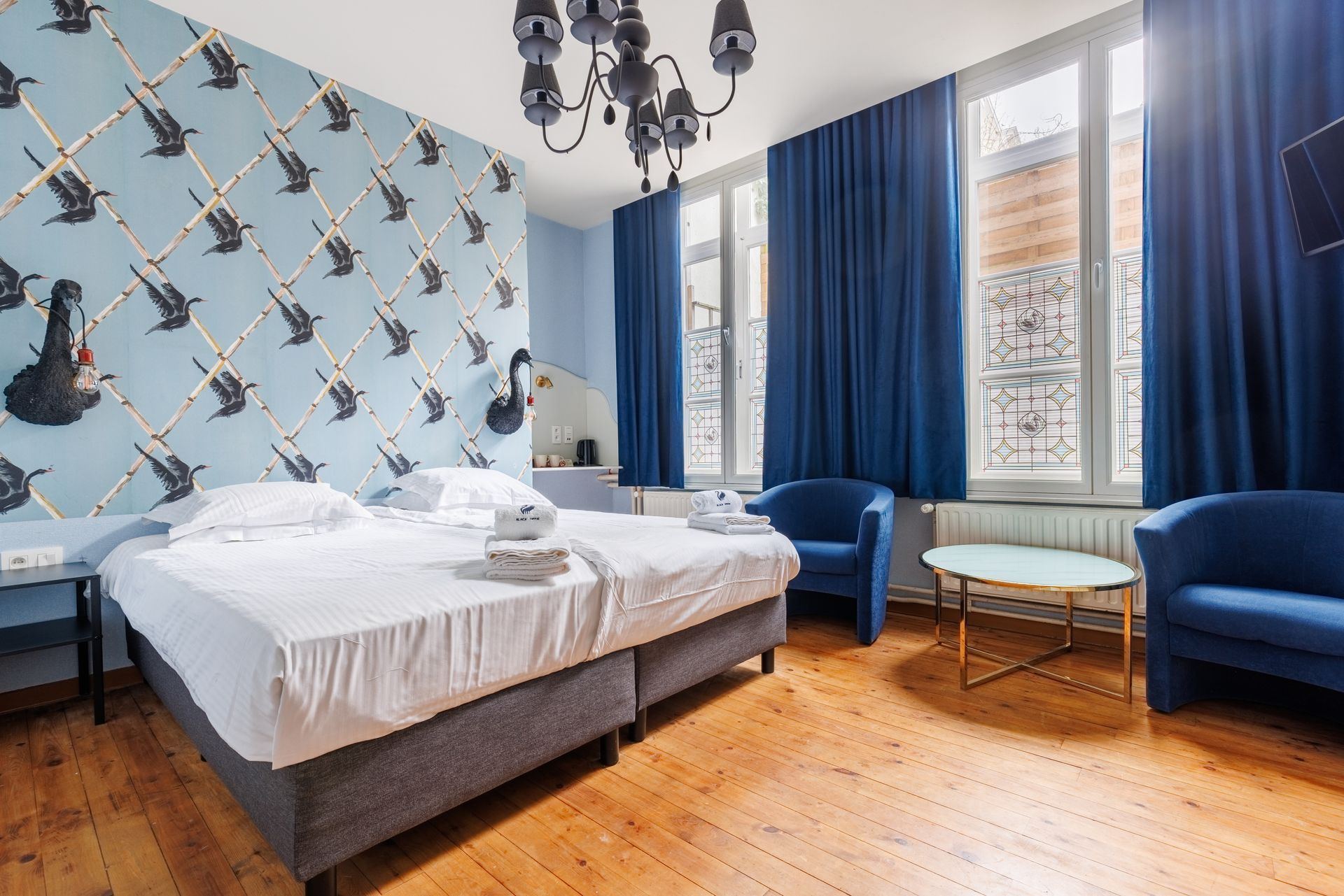 A hotel room with two beds, light blue bird-patterned wallpaper, a black chandelier, and two navy blue armchairs.