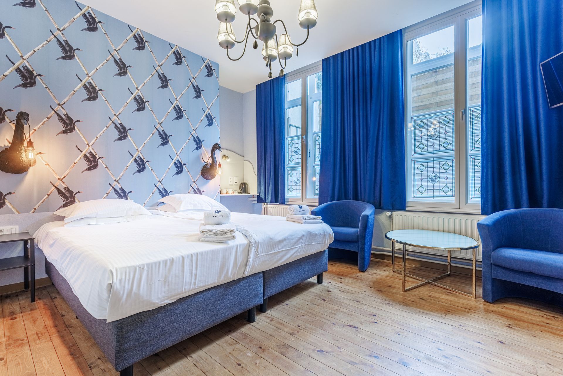 A bedroom with two blue armchairs, a small table, a double bed, and walls decorated with blue bird-patterned wallpaper.
