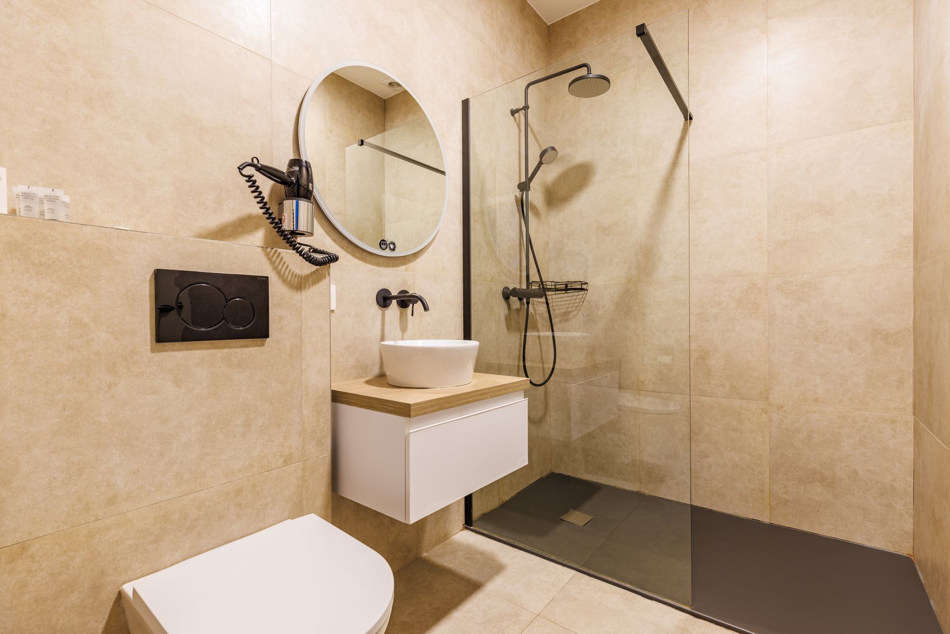 Modern bathroom with beige tiled walls, a white sink on a wood vanity, a round mirror, a toilet, and a glass-walled shower.