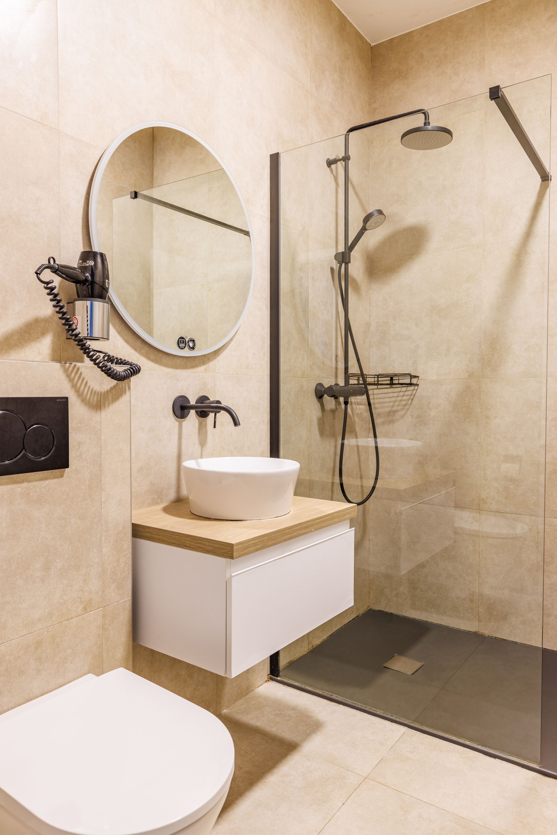 A modern bathroom with a white vessel sink, floating vanity, circular mirror, and a glass-enclosed shower with dark tiles.
