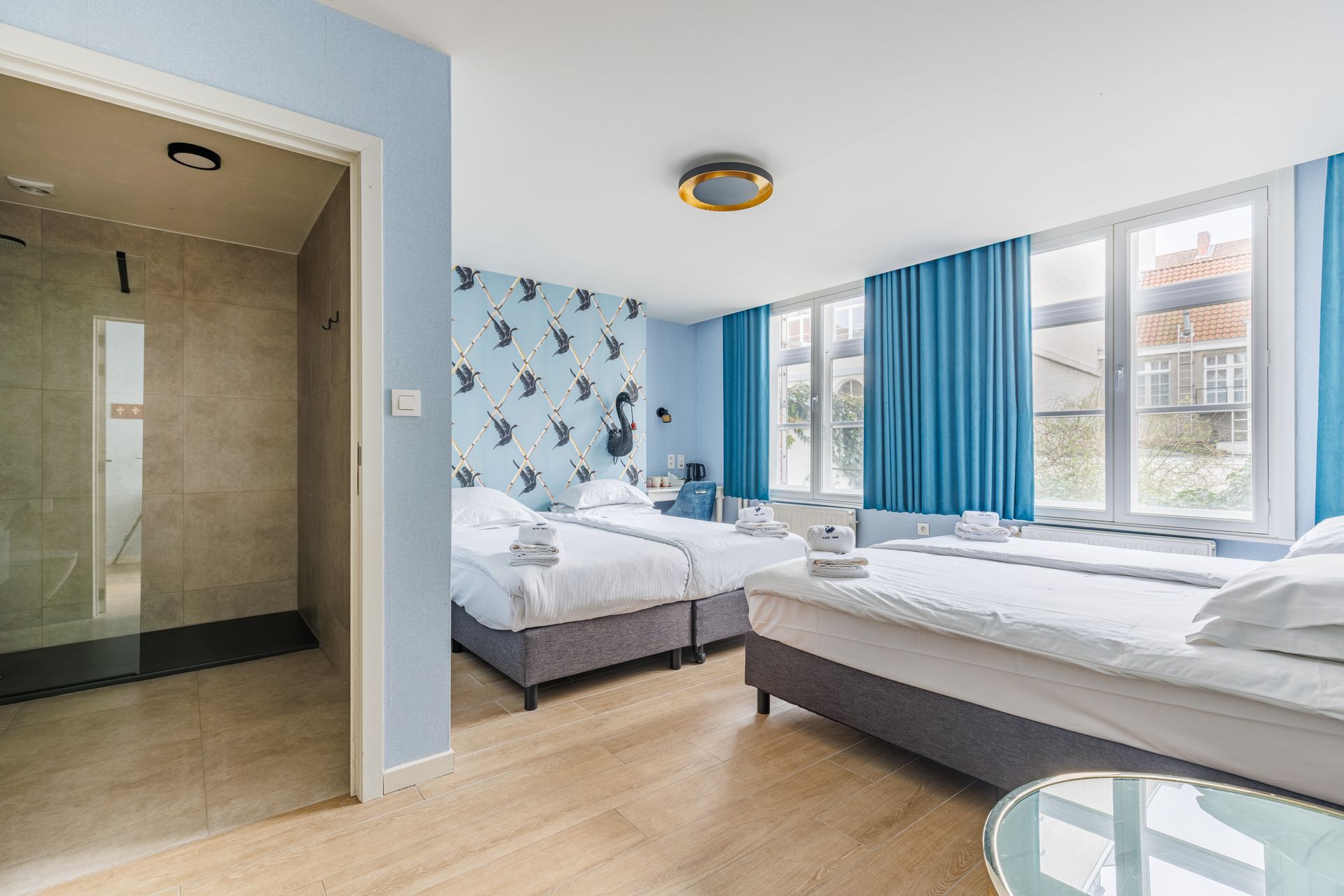 A hotel room with two white beds, blue patterned wallpaper, and an open en-suite bathroom with a glass shower stall.