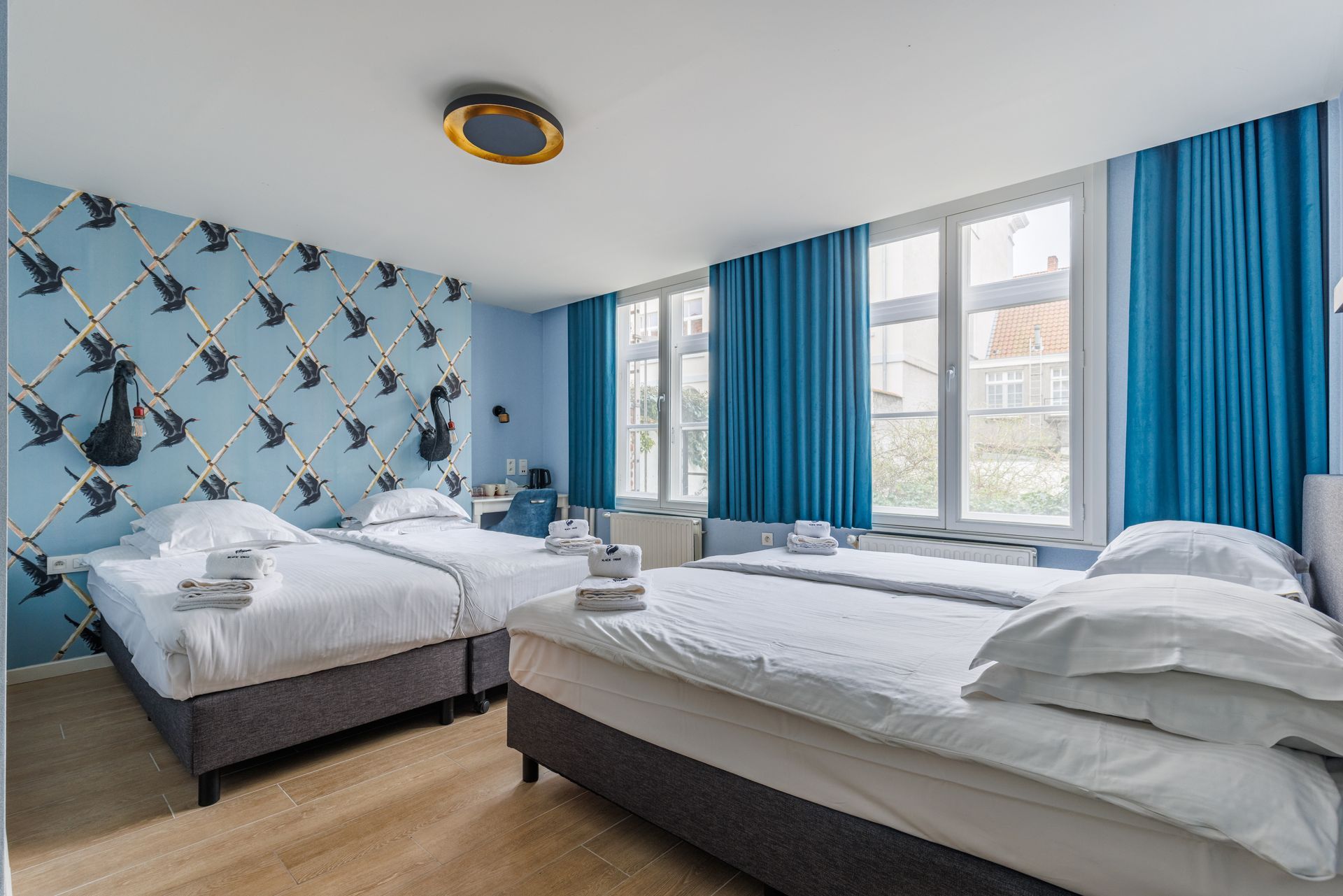 Hotel room with two twin beds, blue swan-patterned wallpaper, and floor-to-ceiling blue curtains.