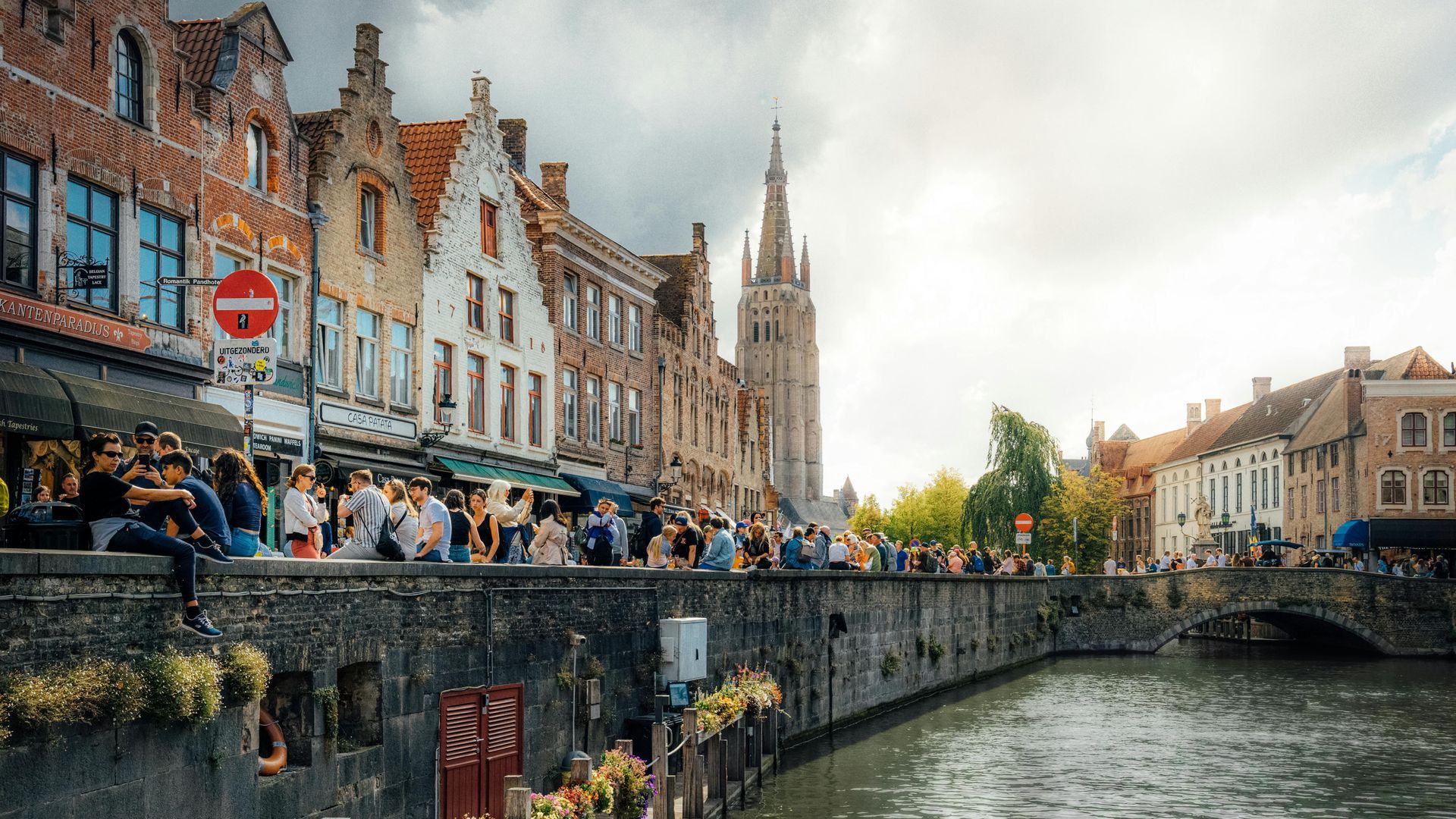A canal scene in Bruges, Belgium, featuring historic brick buildings, a prominent church spire, and a crowd by the water.