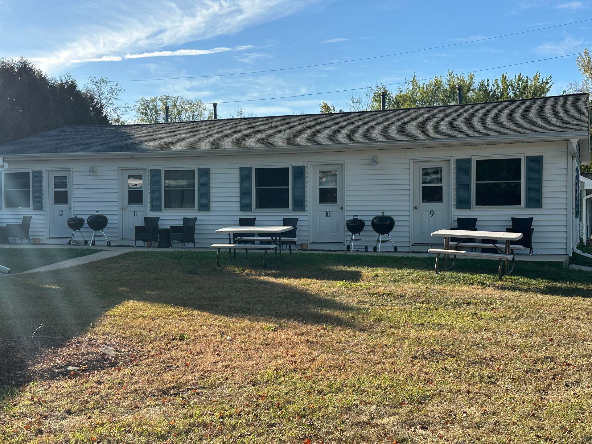 Row of white buildings with doors, windows, and picnic tables. Blue shutters, grills, and green grass.