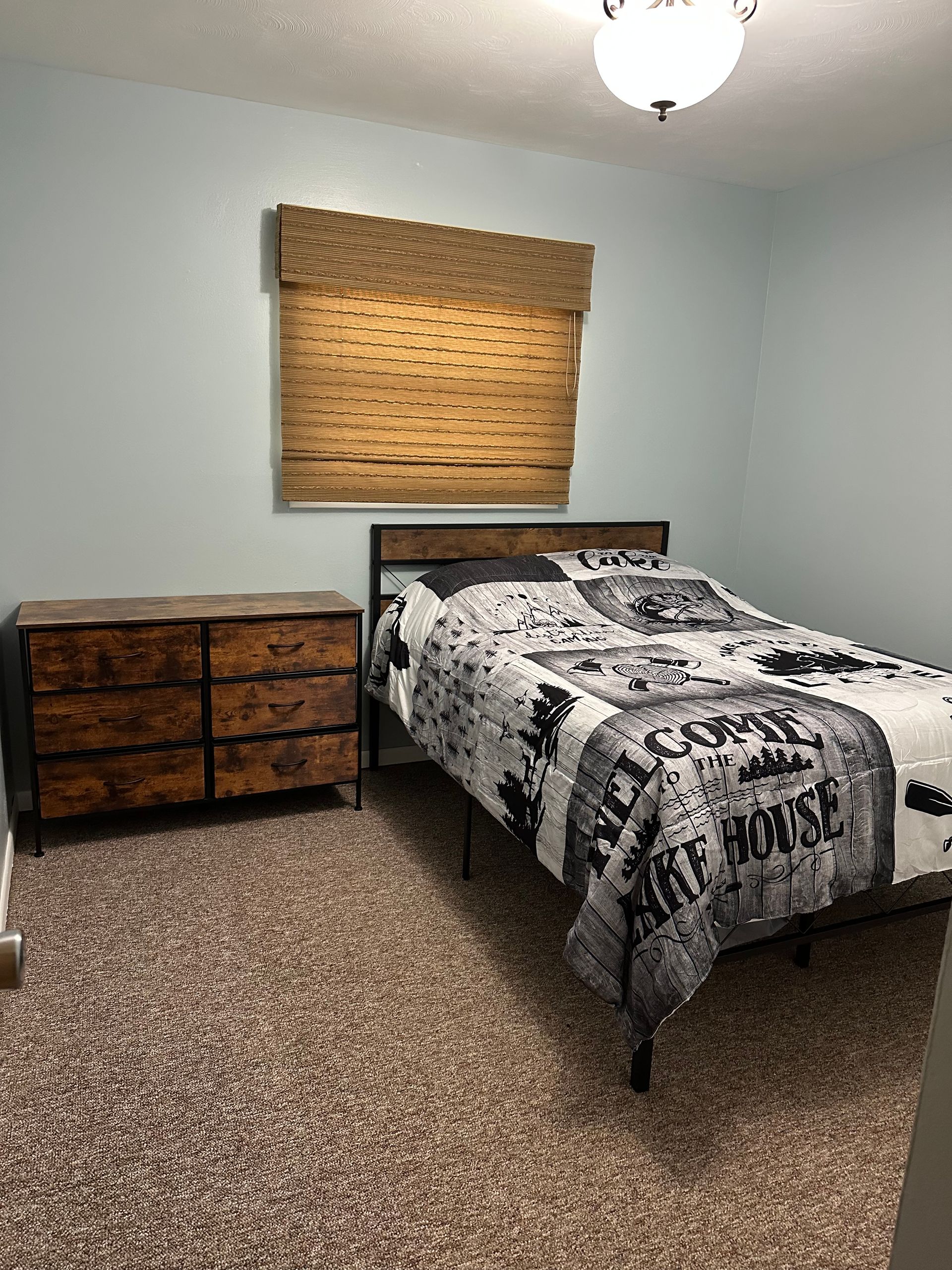 Bedroom with dresser, bed, and window with bamboo shade, tan carpet and light blue walls.