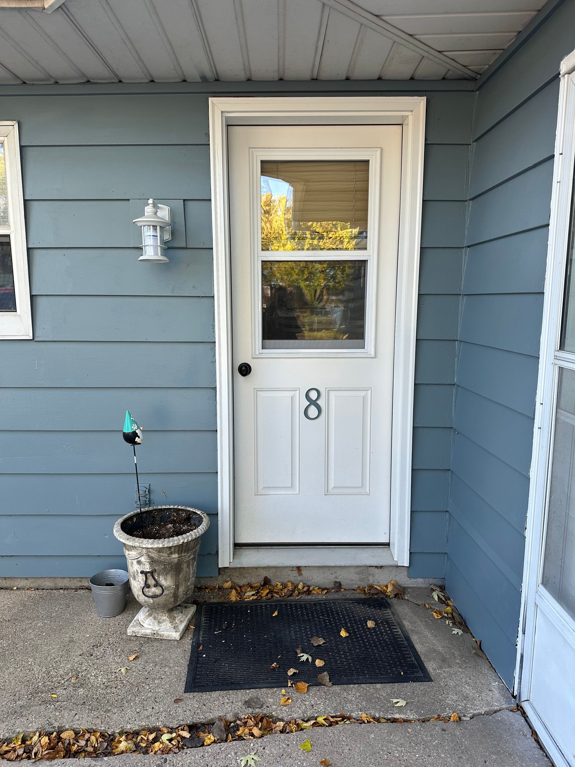 White door with window, number 8, blue siding, small porch with welcome mat and planter.