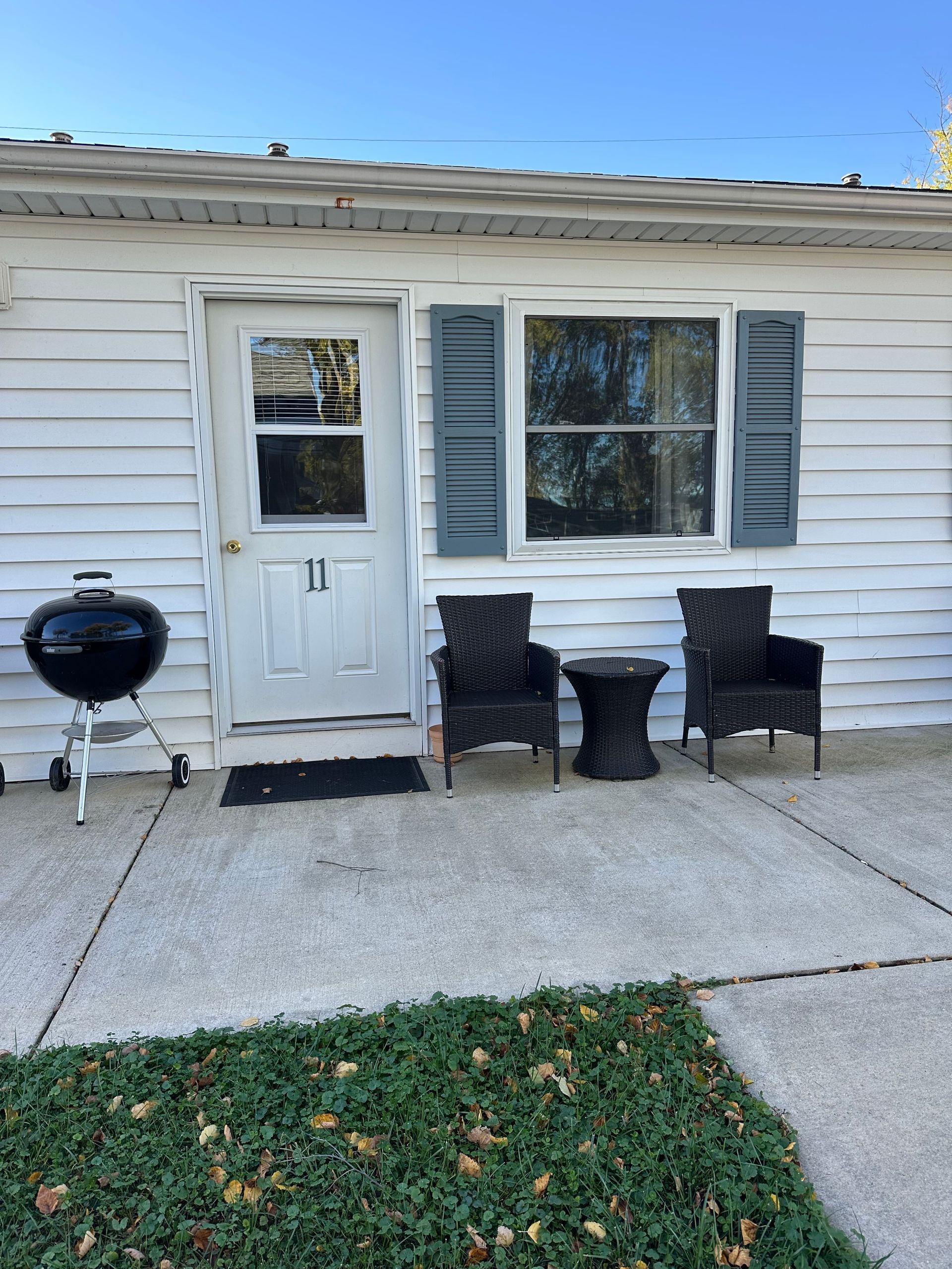 White cottage exterior with door, window, two chairs, small table, grill, and blue shutters.