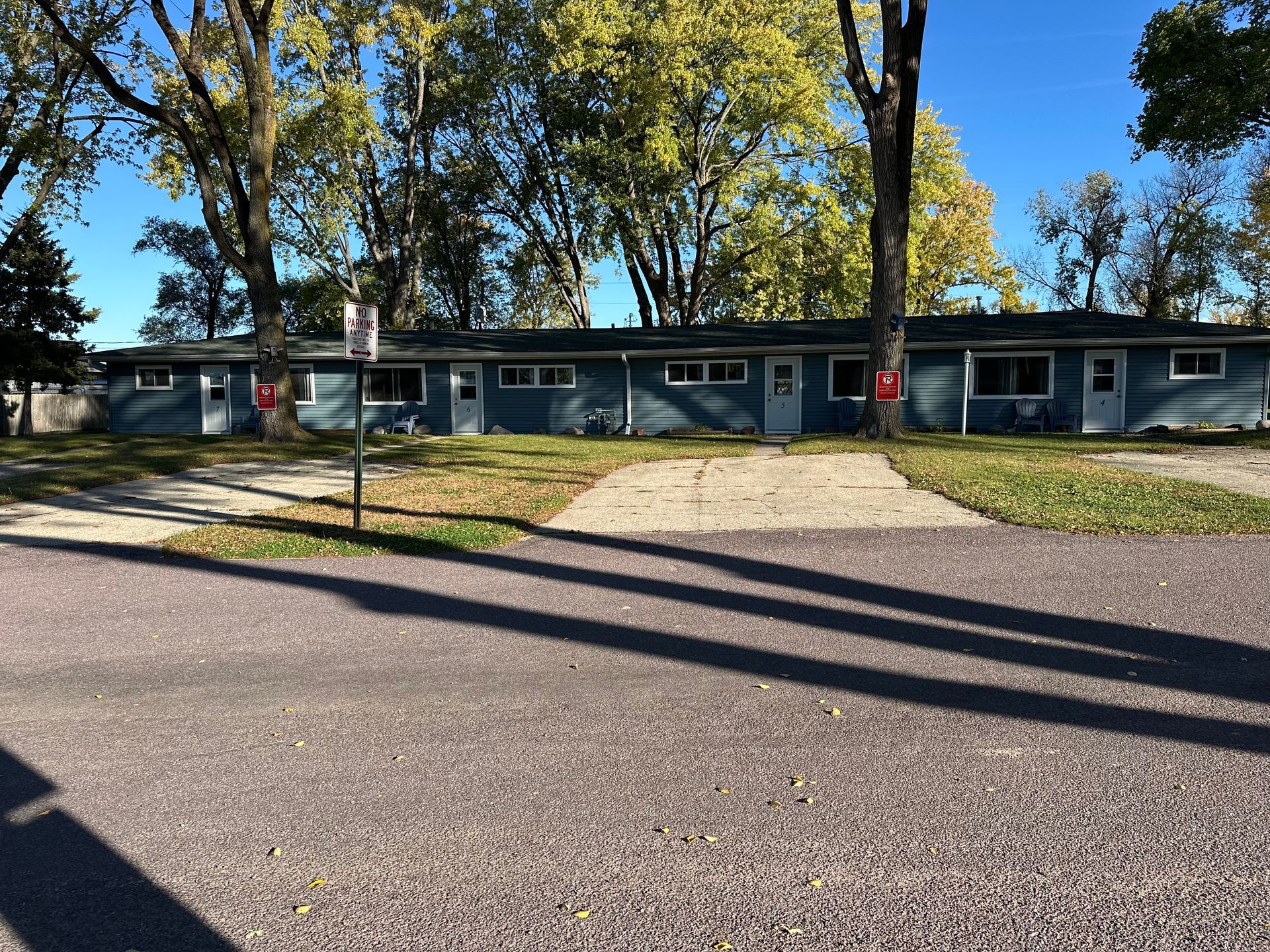 A row of blue single-story apartments with white doors and windows. Trees flank the building, casting long shadows.