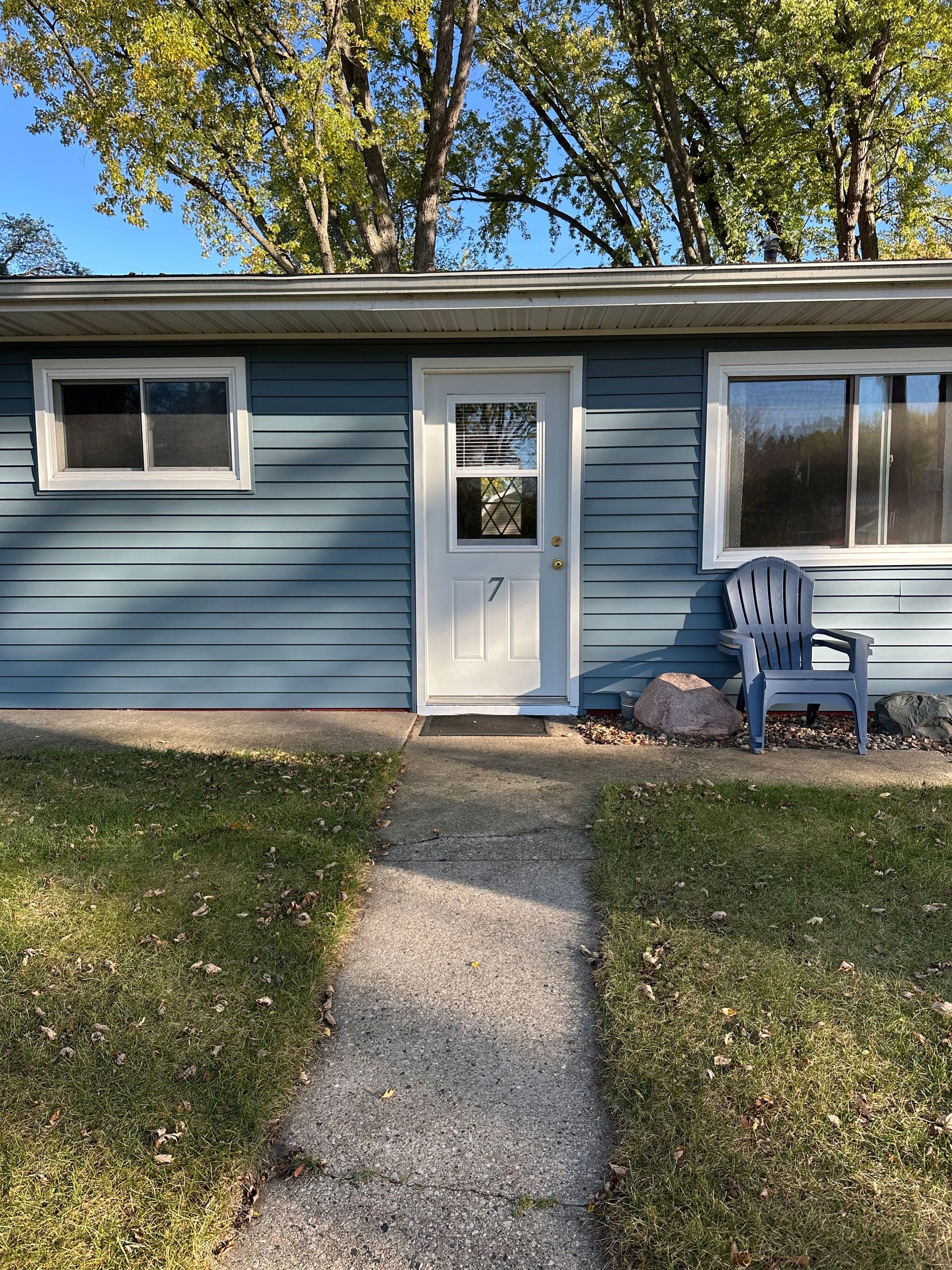 Blue house with a white door, windows, and a path leading to it. A blue chair sits beside a rock.