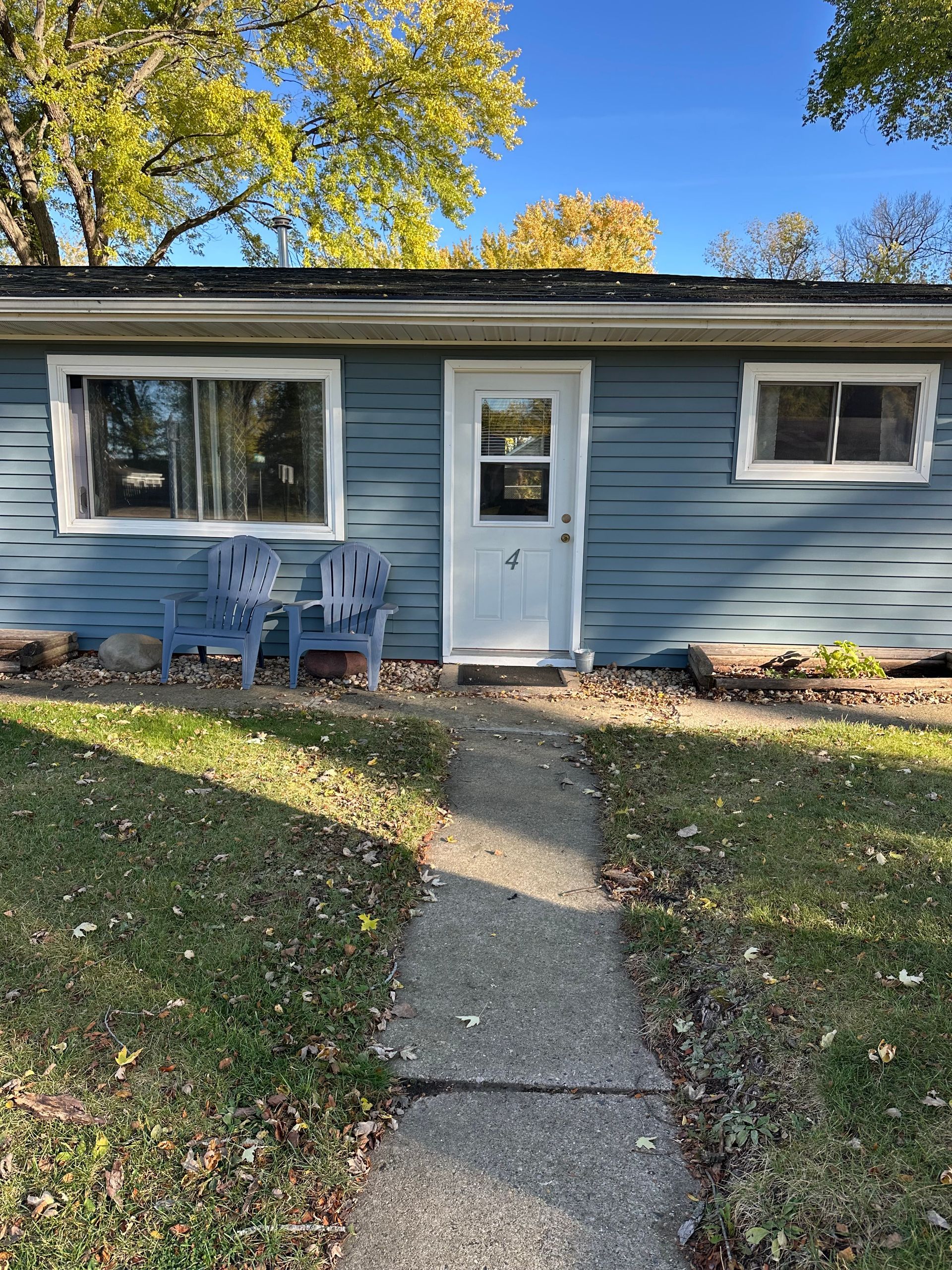 Blue house with white door and windows, concrete path, two blue chairs on lawn, autumn trees.