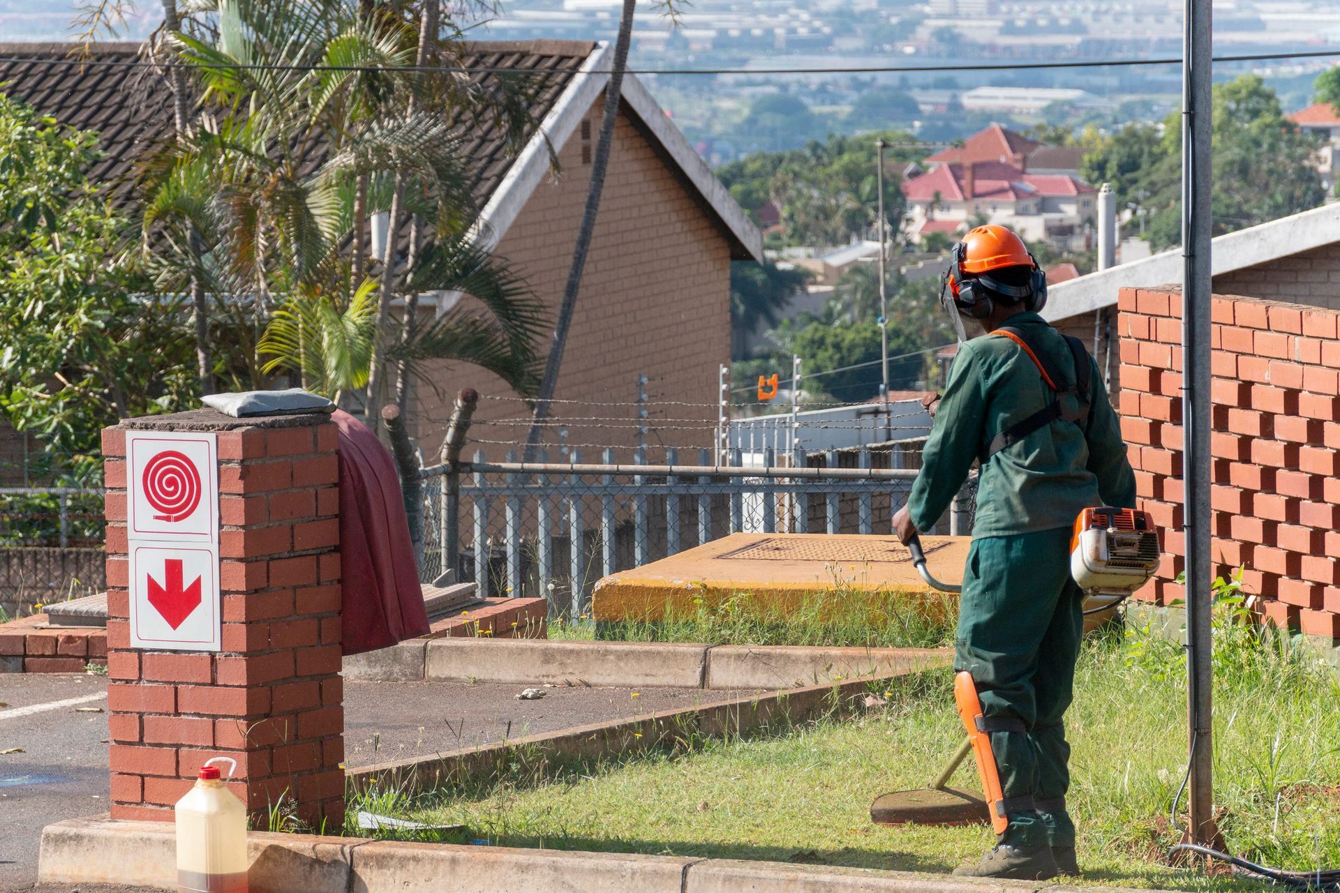 A worker in green uniform and safety gear uses a weed trimmer to clear grass next to a brick pillar with a fire sign.