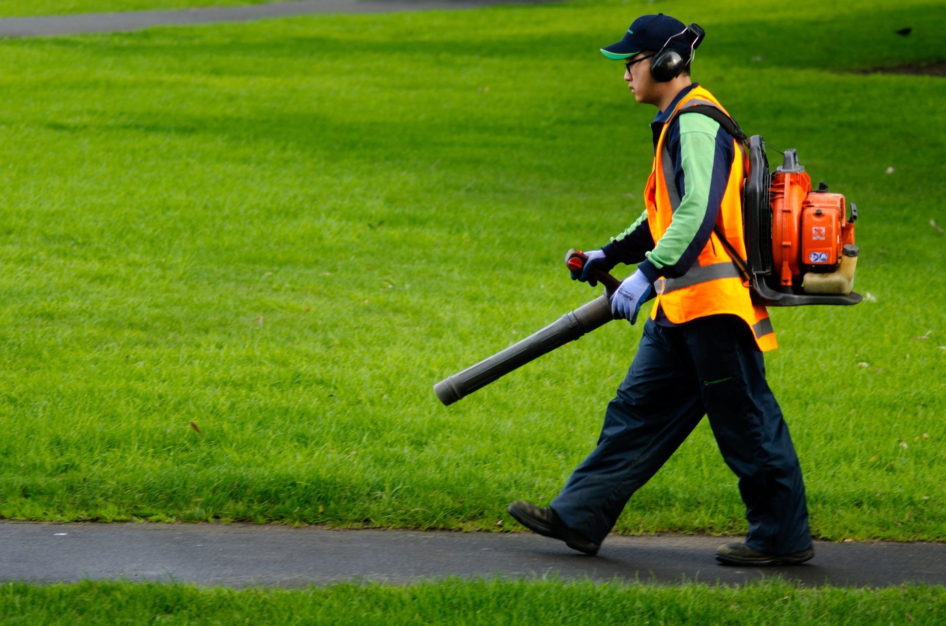 A worker in a high-visibility orange vest and ear protection walks along a path, using a backpack leaf blower.