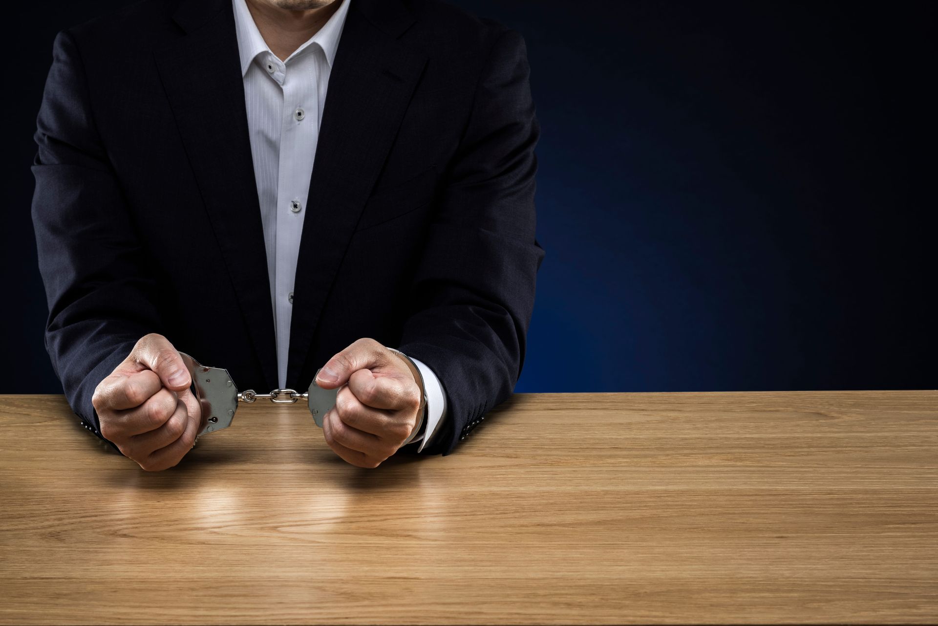 A person in a dark suit with hands in metal handcuffs resting on a wooden table against a dark blue background.