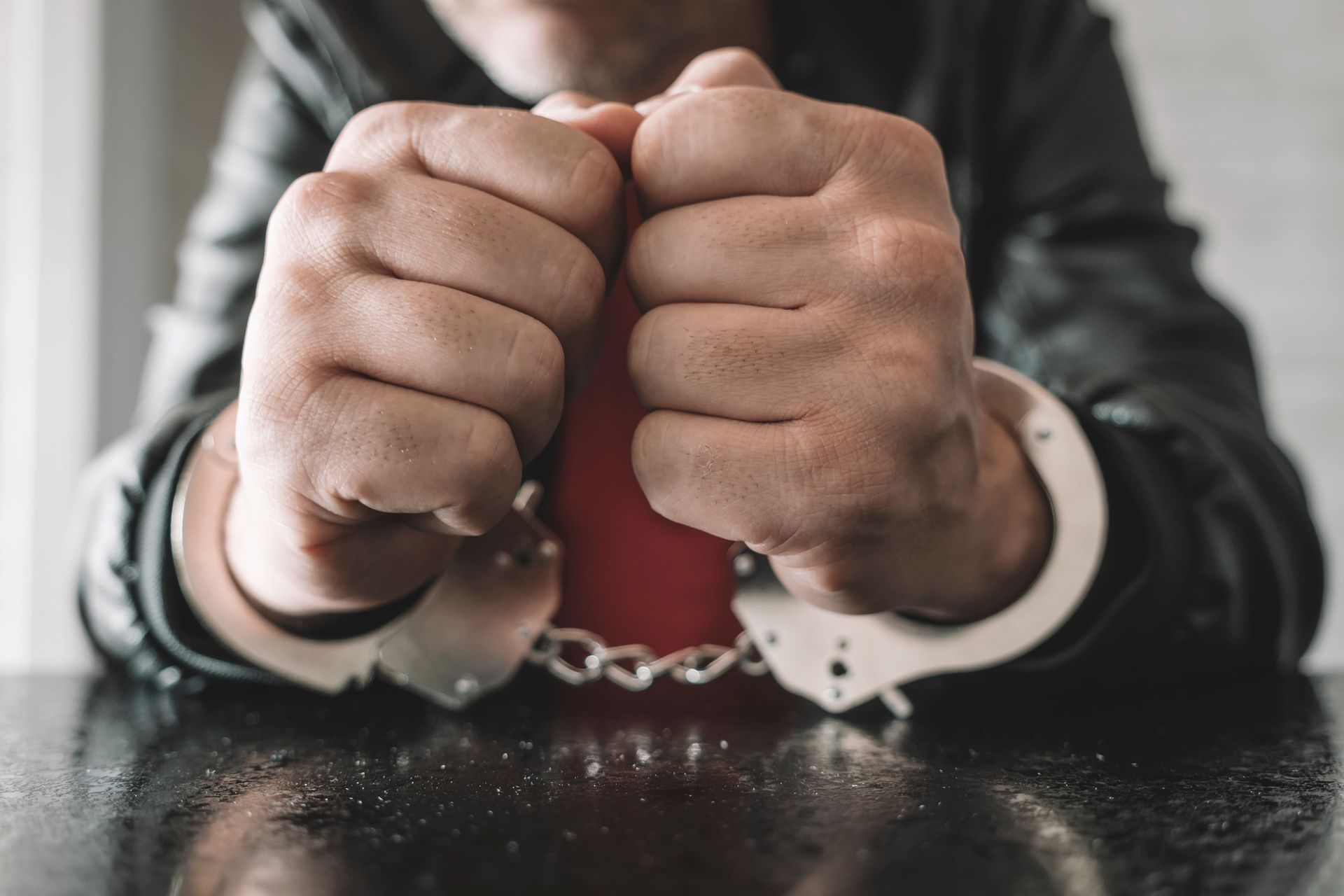 Handcuffed hands clenched on a table, with a blurred person in the background.