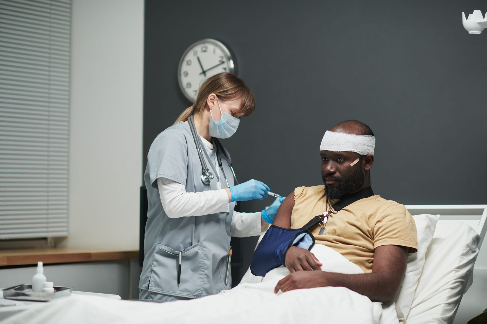 A medical professional wearing a mask and scrubs administers an injection into a patient’s arm in a hospital room.