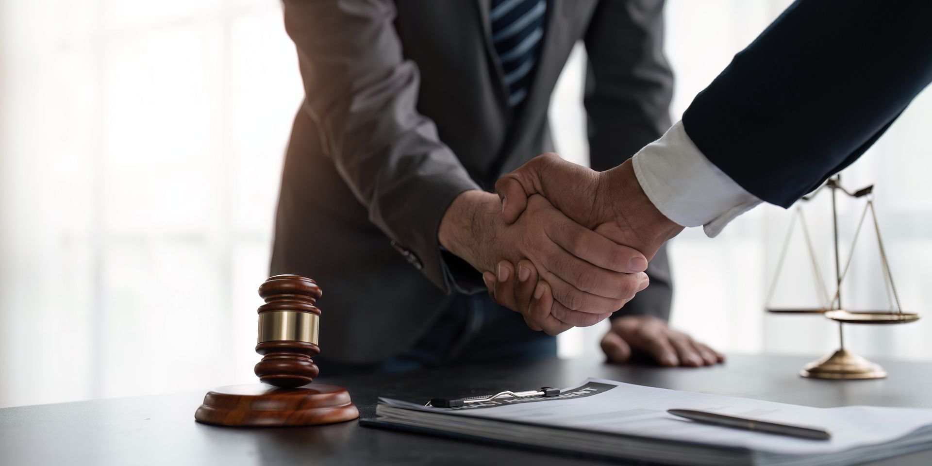 Two people in suits shaking hands at a desk with a gavel, scales of justice, and documents.