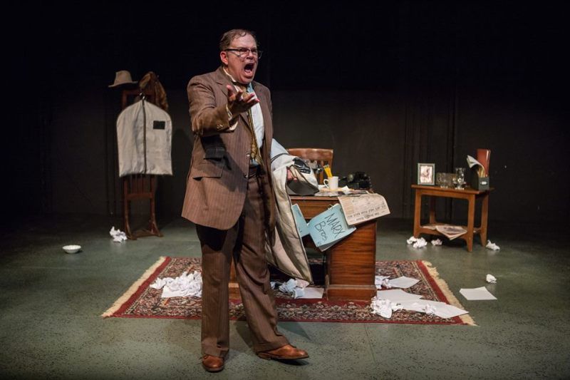 Bernie standing in front of desk yelling while holding garment bag and gesturing.