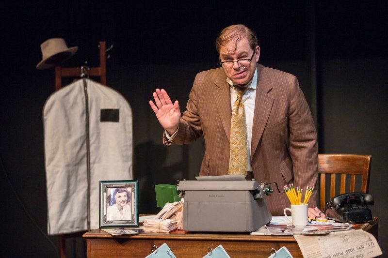 Bernie, grimacing, standing behind desk with right arm raised.