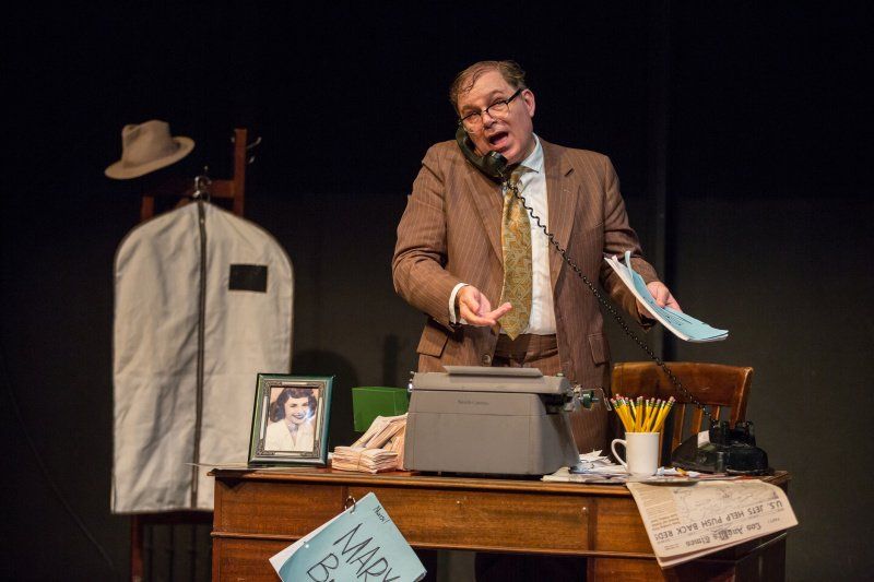 Bernie standing behind messy desk talking on phone and holding a script.