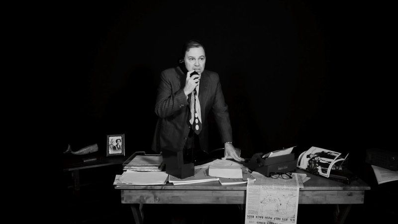 Bernie standing behind his desk on phone he's holding in right hand.