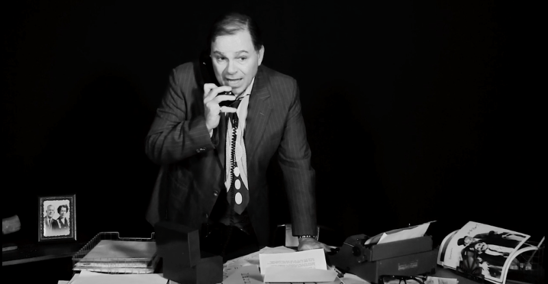 Bernie standing behind desk holding phone in right hand while on a call.