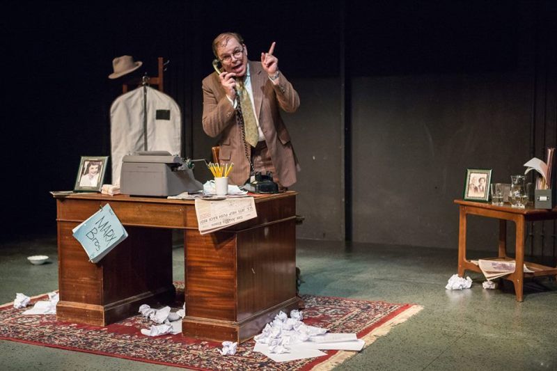 Bernie standing behind messy desk holding  phone in right hand and raising left hand.