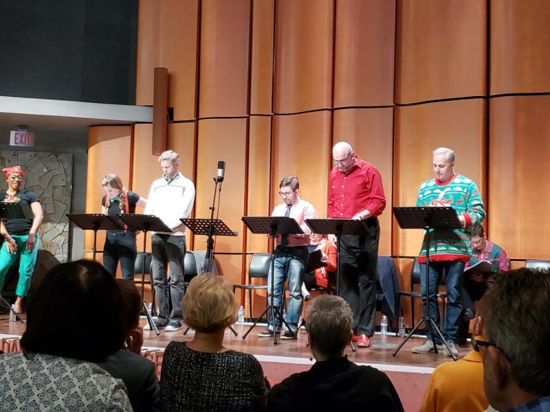 1 actor in green pants and nightcap listens while 5 actors standing at music stands continue a scene
