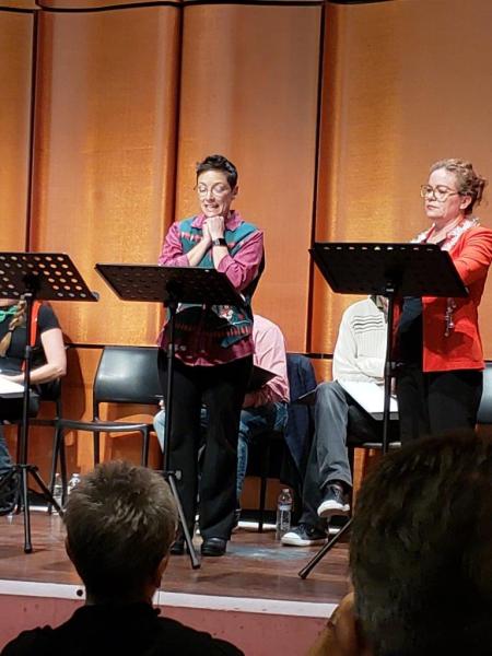 Woman in pink blouse and sweater vest talking to woman in red jacket during a scene