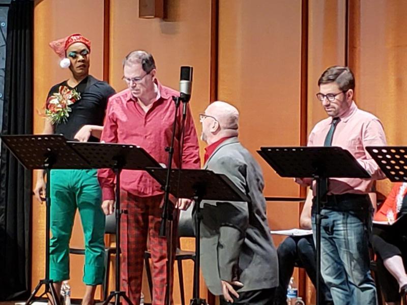 3 actors standing at music stands and one in grey jacket kneeling