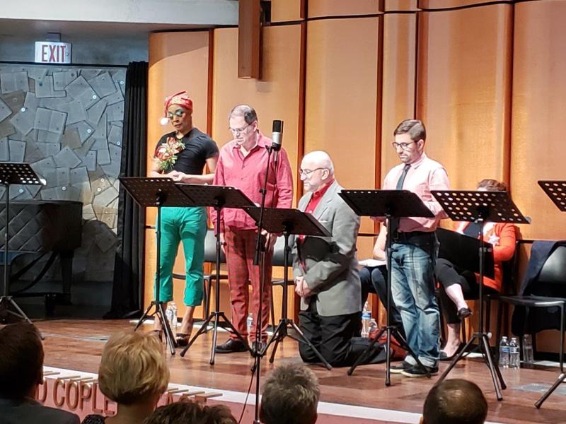 4 actors at music stands with one in grey jacket kneeling