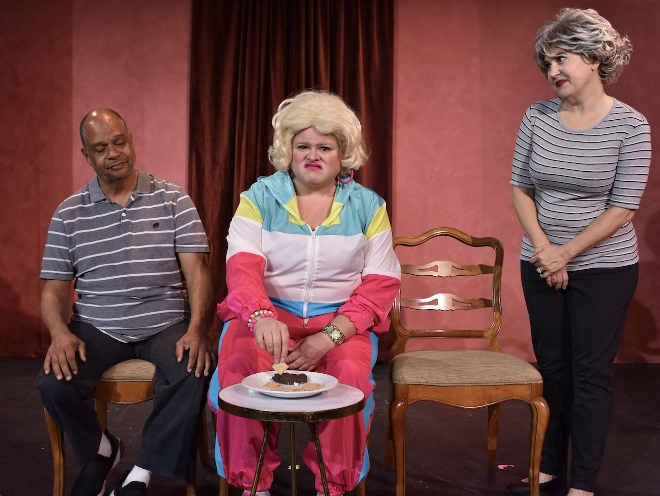 A woman in a wig is sitting with two other people at a small table with a plate of food on it.