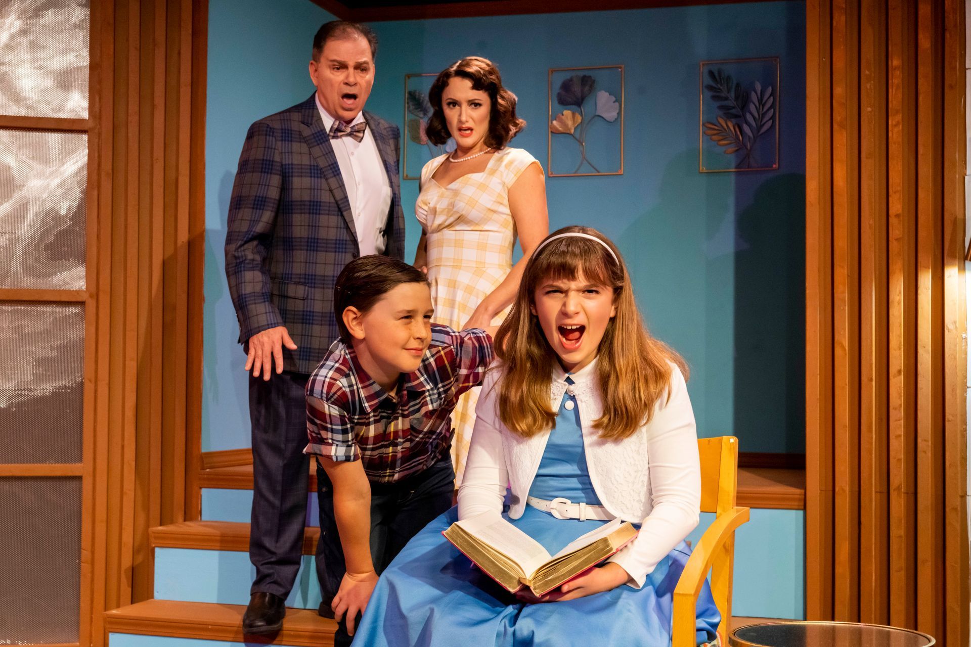 A group of people standing around a girl sitting in a chair holding a book .