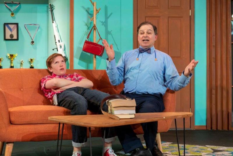 Father in blue shirt and son in red-checked shirt having a conversation on orange sofa.