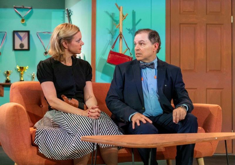 Mother in black blouse and gun belt and father in suit jacket talking while seated on sofa.