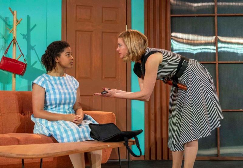 Mother in black and white striped dress giving gun to daughter in blue and white dress on sofa.
