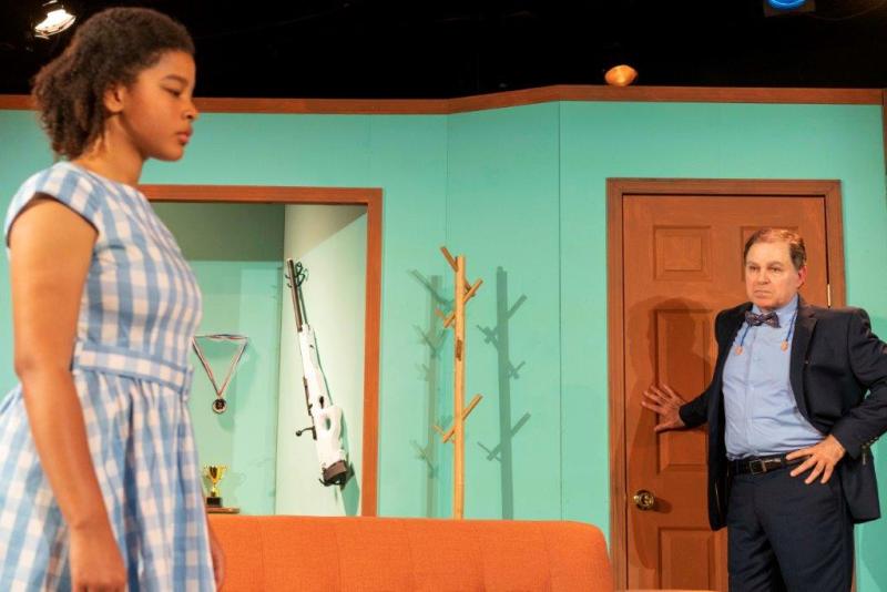 Father in grey standing at door looking at daughter in blue and white dress near sofa.