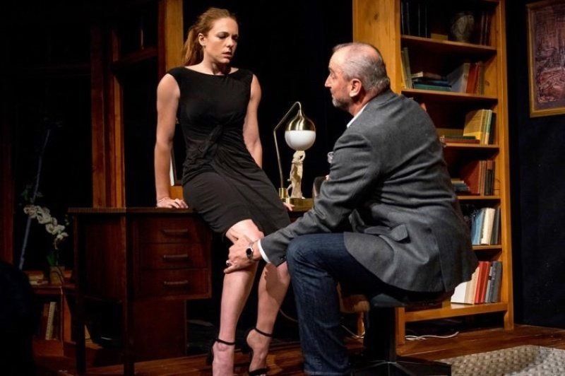 Young woman in black dress seated on piano with seated man in grey jacket caressing her right leg