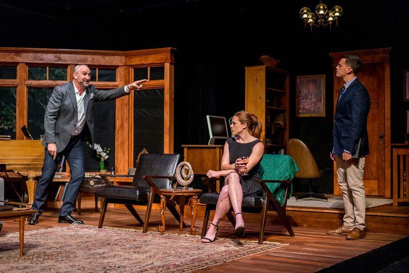 Man in grey suit smiling and gesturing as woman in black dress and man in blue suit jacket  watch