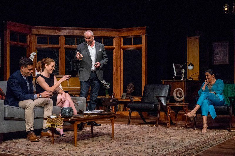 Couple on sofa drinking wine, man in grey suit smiling and gesturing as seated woman in blue watches