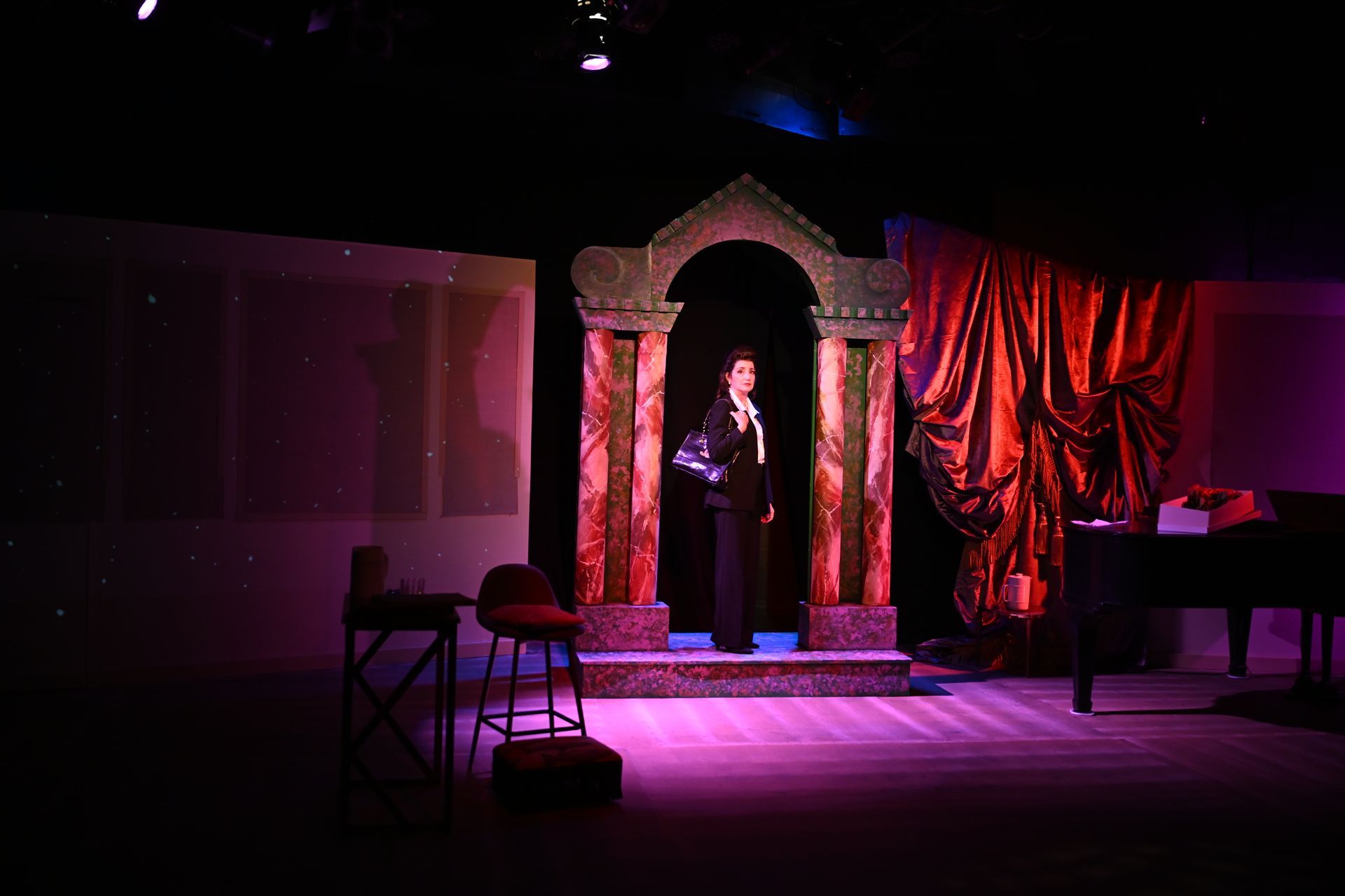 A person stands on a stage, framed by a decorated archway. Red curtains and a piano are visible.