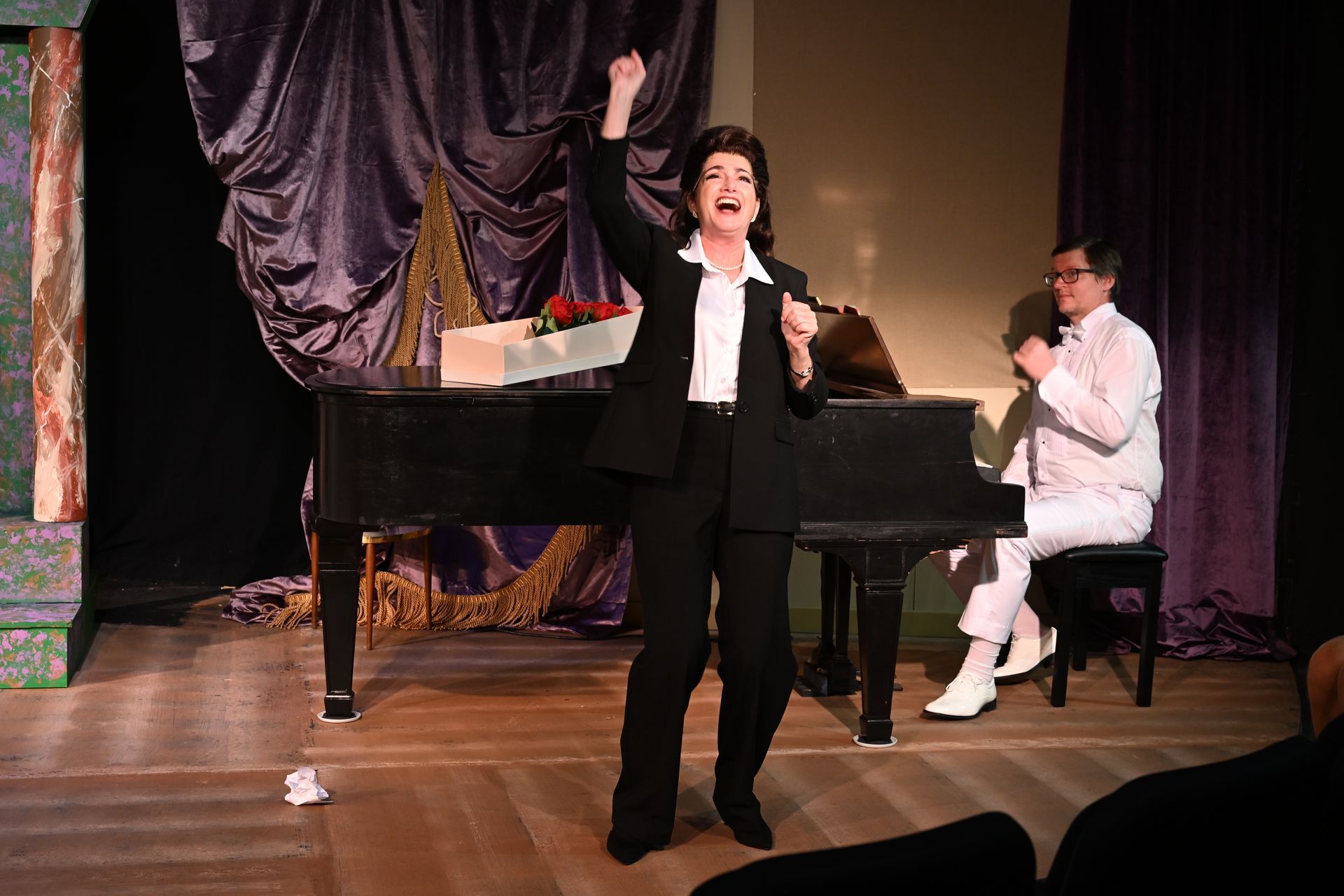 Woman in a black suit celebrates on stage with her fist in the air. A man in white plays piano.