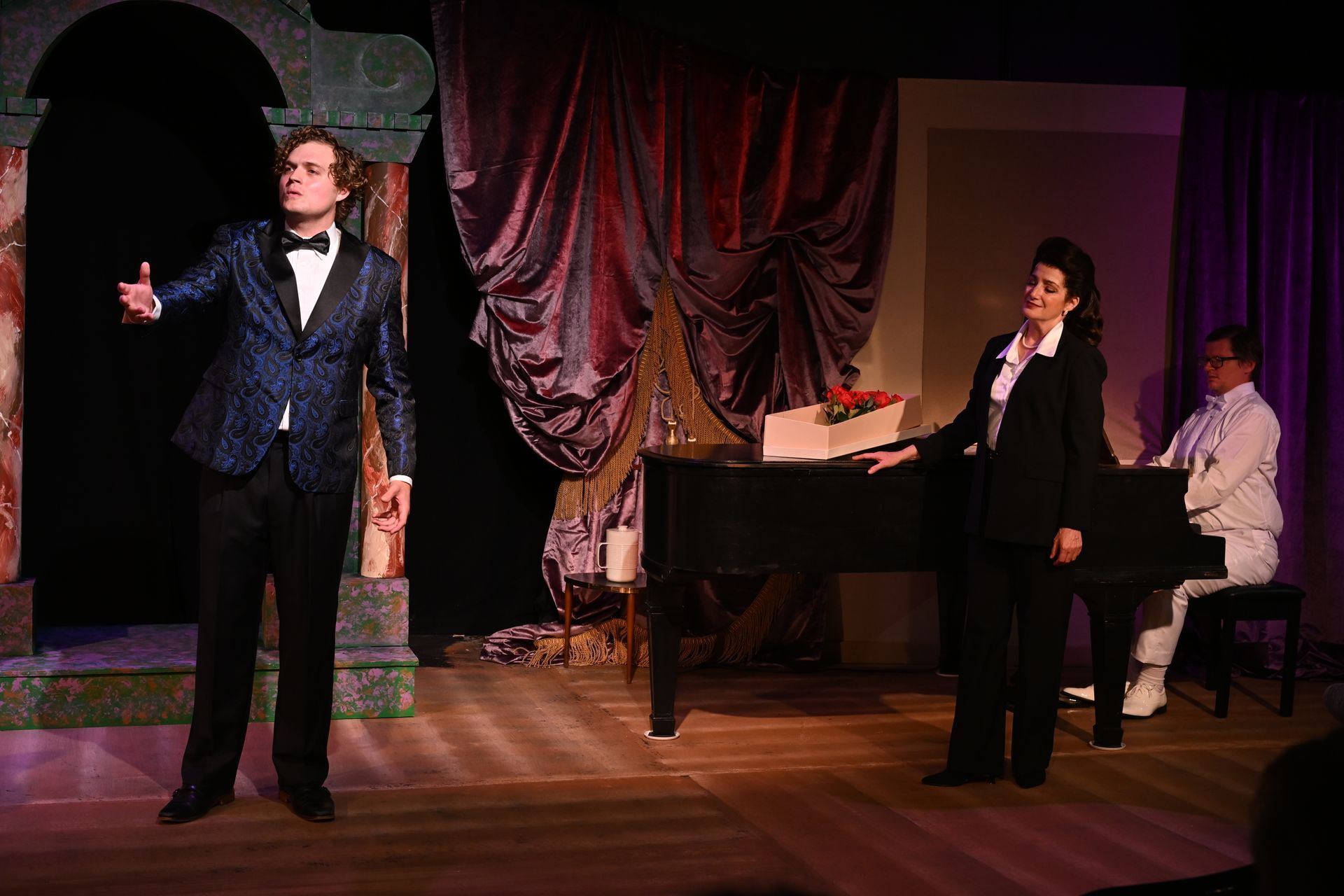 Man in blue suit sings on stage, woman in black suit standing beside a piano. Pianist sits at piano stage.