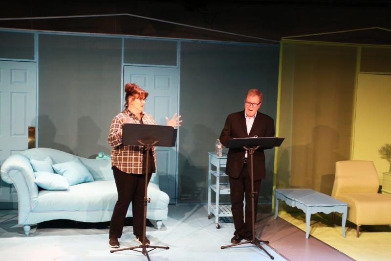 Woman in brown checked shirt and pants and man in brown suit onstage during play reading.