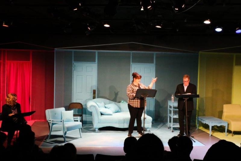 Woman in brown checked shirt in front of blue sofa gesturing at man in brown suit.