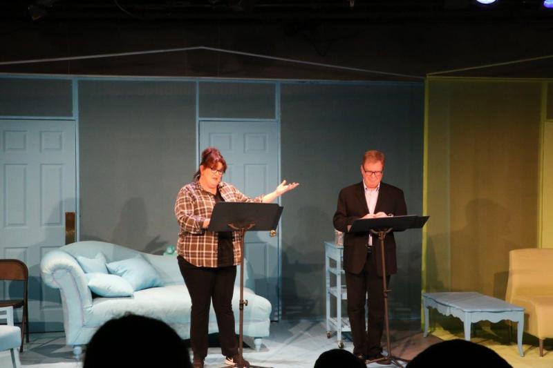 Woman in brown checked shirt with left arm up and man in brown suit listening to her.