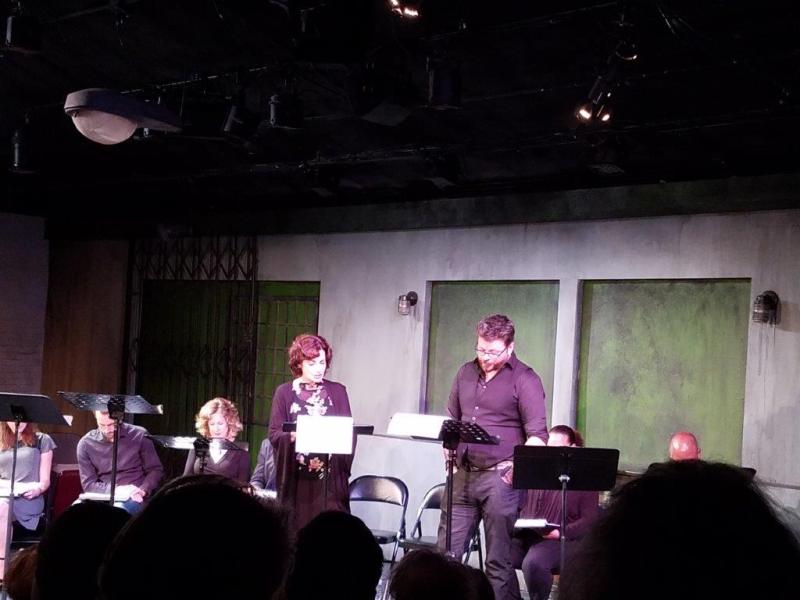 Play reading featuring many actors standing at music stands or seated.