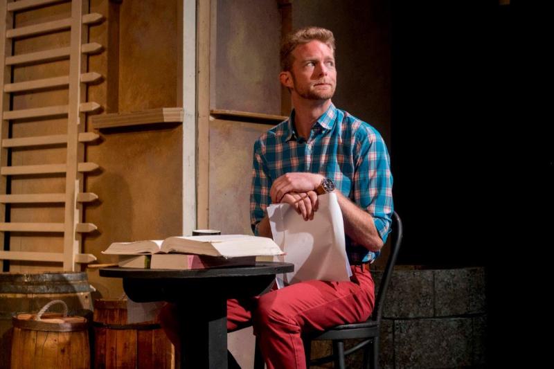Actor Brian Mackey sitting at a desk looking up while holding a script in his hands