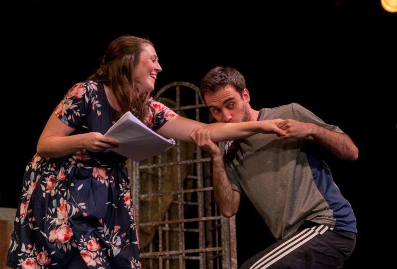 Woman in flowered blouse rehearsing scene with man in grey shirt kissing her left arm.