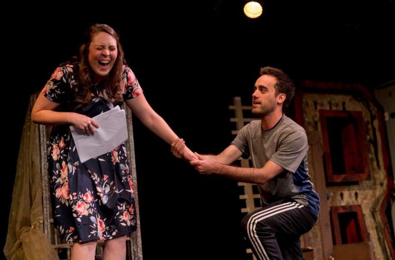 Woman in flowered dress laughing with kneeling man in grey while rehearsing.