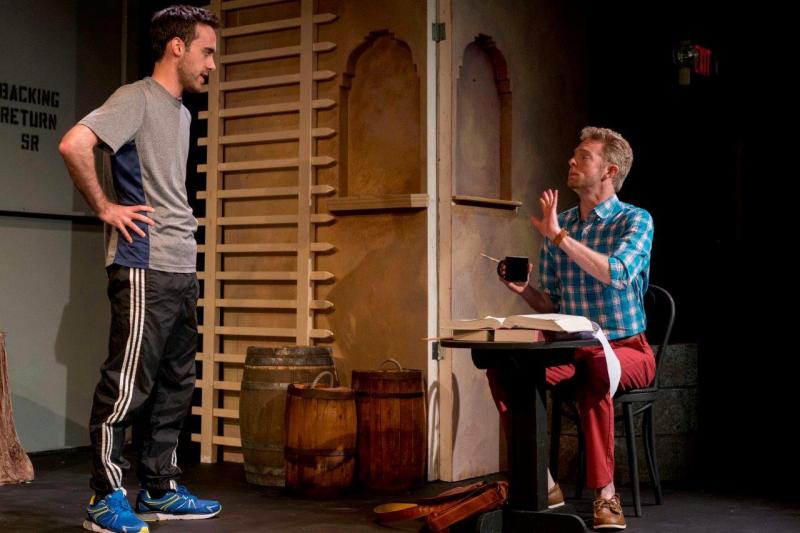 Man in grey shirt talking with man in blue checked shirt sitting at a desk and holding coffee cup.
